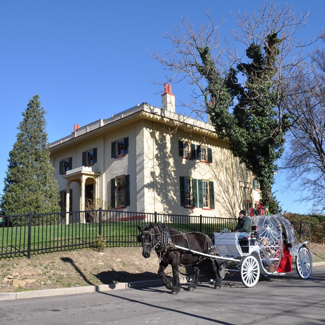 Horse drawn carriage riding past a two-story house on top of a hill