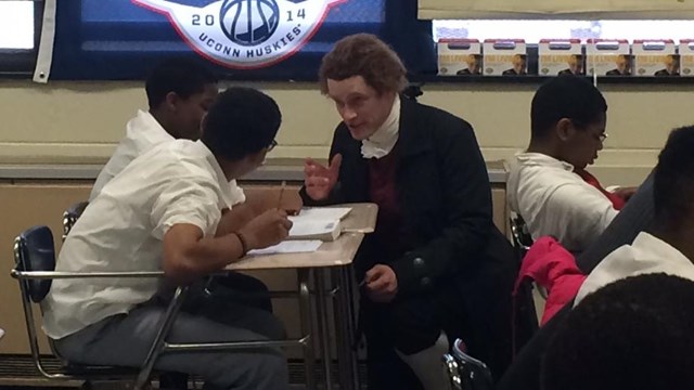 Color photo showing a man dressed in colonial costume talking with students who are seated at desks.