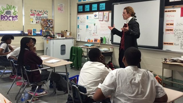 Color photo of a man in colonial costume standing in front of a class of students in a classroom.
