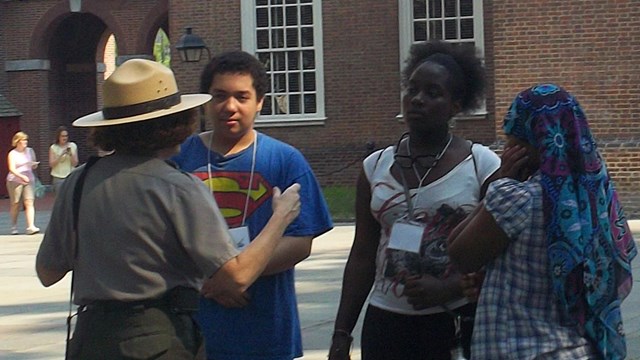 Color photo of a female ranger speaking to a group of teens behind Independence Hall.