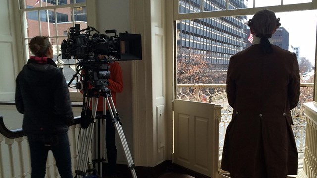 Color photo of a cameraman filming a man in 18th century costume in front of a large window.