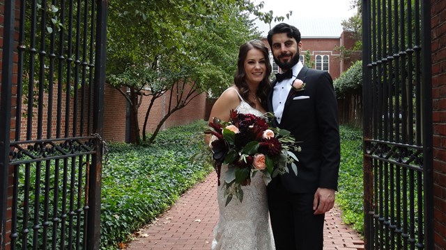 Color photo of a bride and groom posing in front of garden gates with greenery behind them.