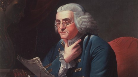 Color photo of a man wearing a white wig and bifocal glasses with his chin resting on his thumb.