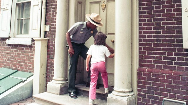 Color photo of a male ranger and a small child standing on a door step, turning a door knob together