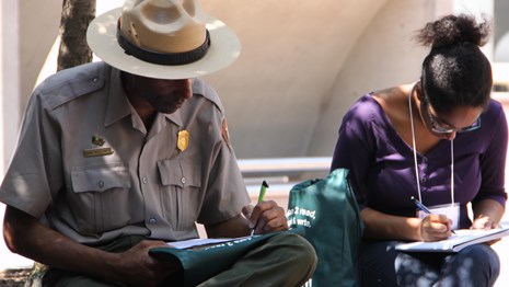Color image showing a male park ranger and a teen girl writing in notebooks in an outdoor setting.