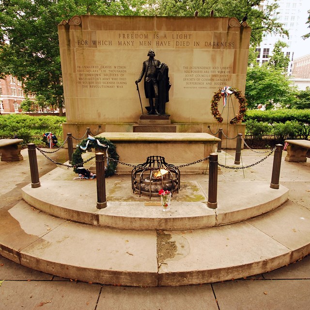 Color photo of a statue of George Washington on a plinth with a tomb and eternal flame below.