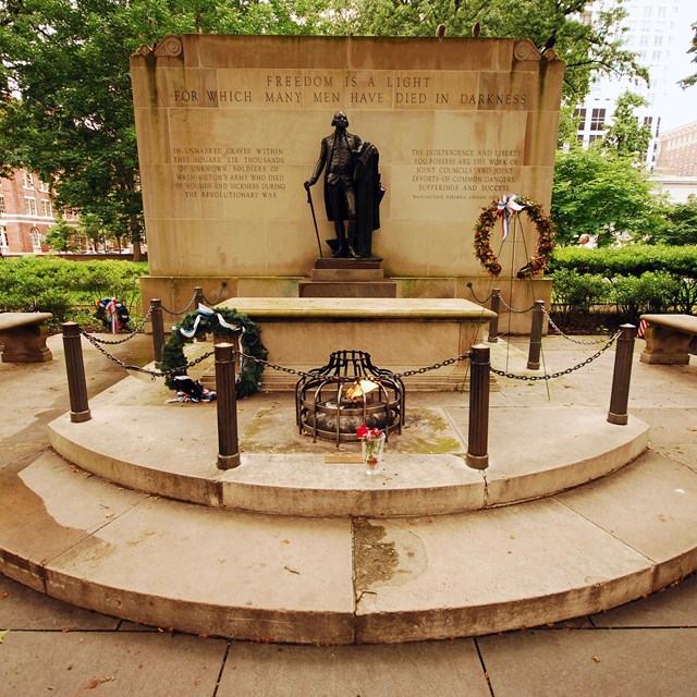 Color photo of a statue of George Washington on a plinth with a tomb and eternal flame below.