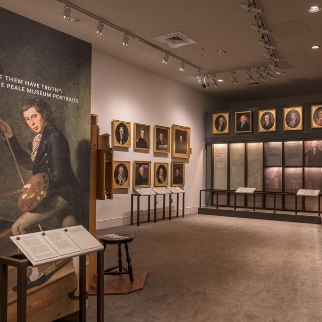 Color photo of exhibit area with a picture of one man (foreground) with rows of portraits behind.