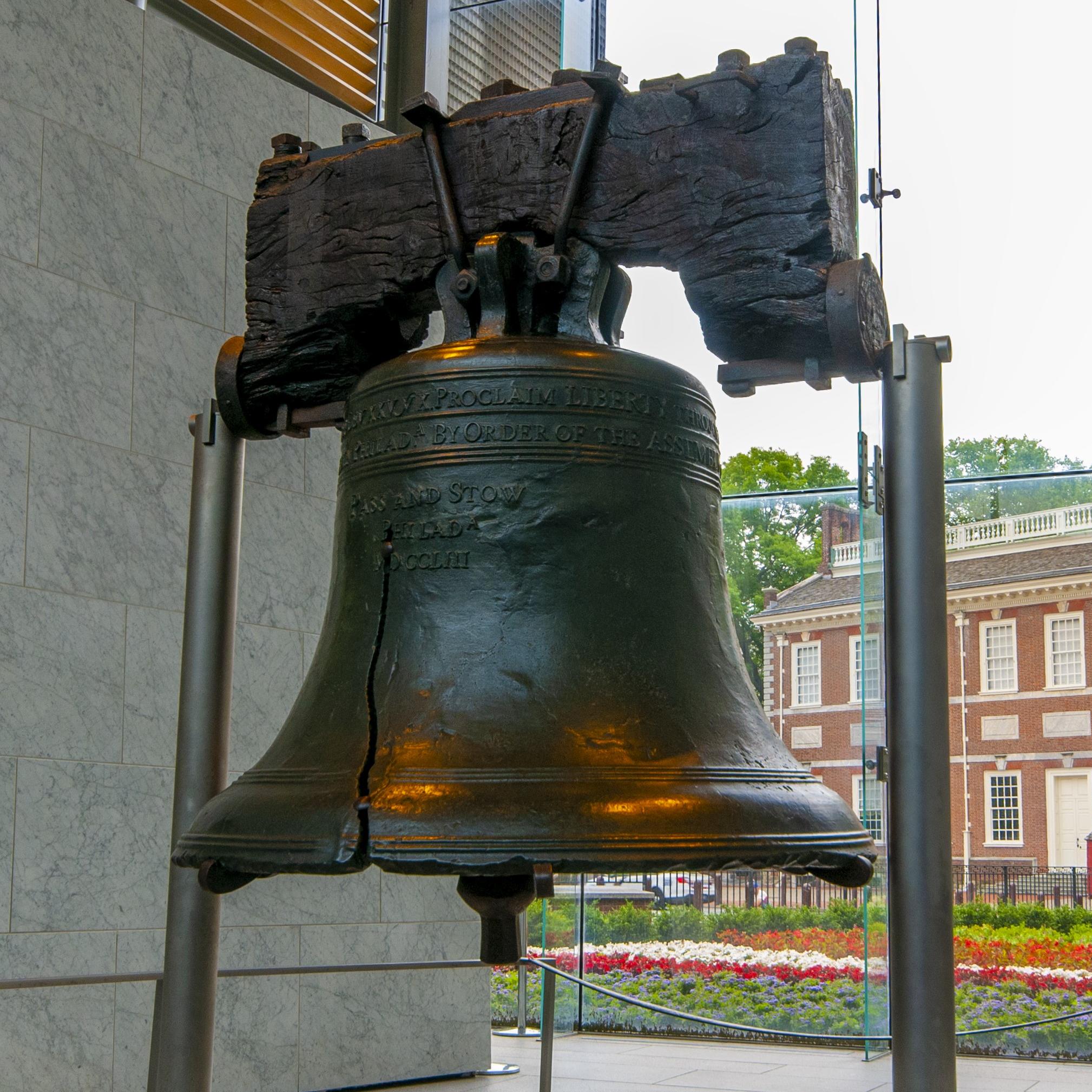 Large suspended bell with a two-story brick building and steeple in the background