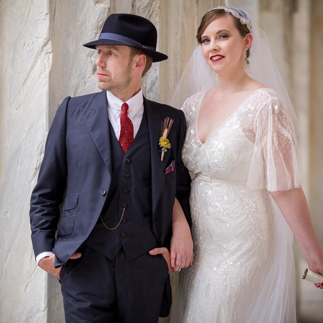 Color photo of a man in a suit and a woman in a wedding dress posing in front of a marble column.