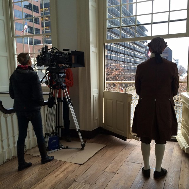 Color photo of a cameraman filming a man in 18th century costume in front of a large window.