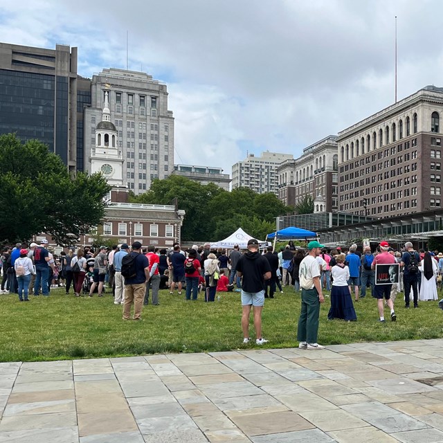 A large group of people, some with signs and some police with bikes, stand facing Independence Hall.