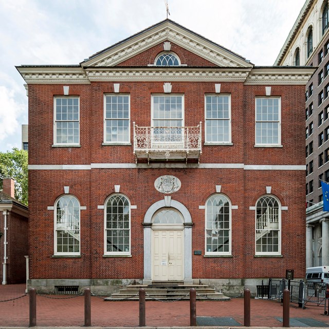 Color photo of the exterior of a two story red brick building with center door and pediment.