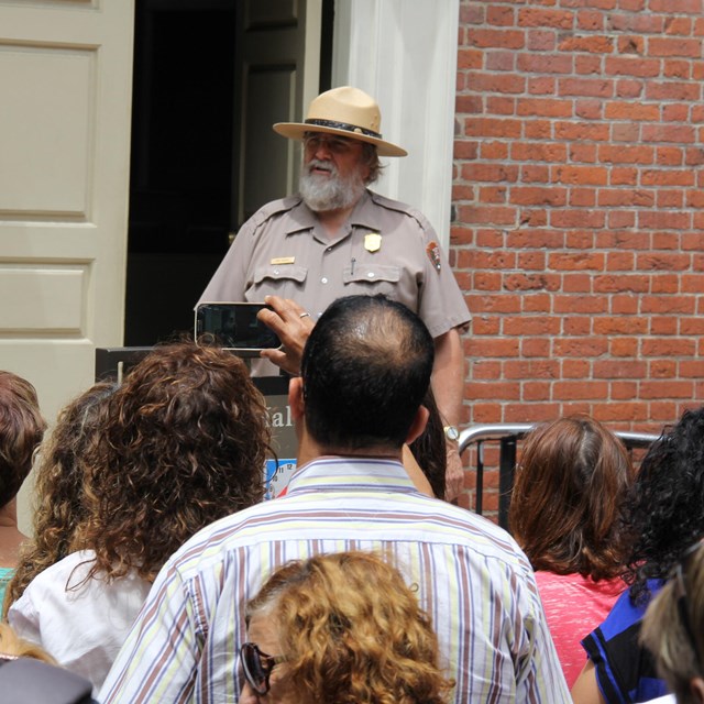 Color photo of a male park ranger standing between an open door and a crowd of visitors.
