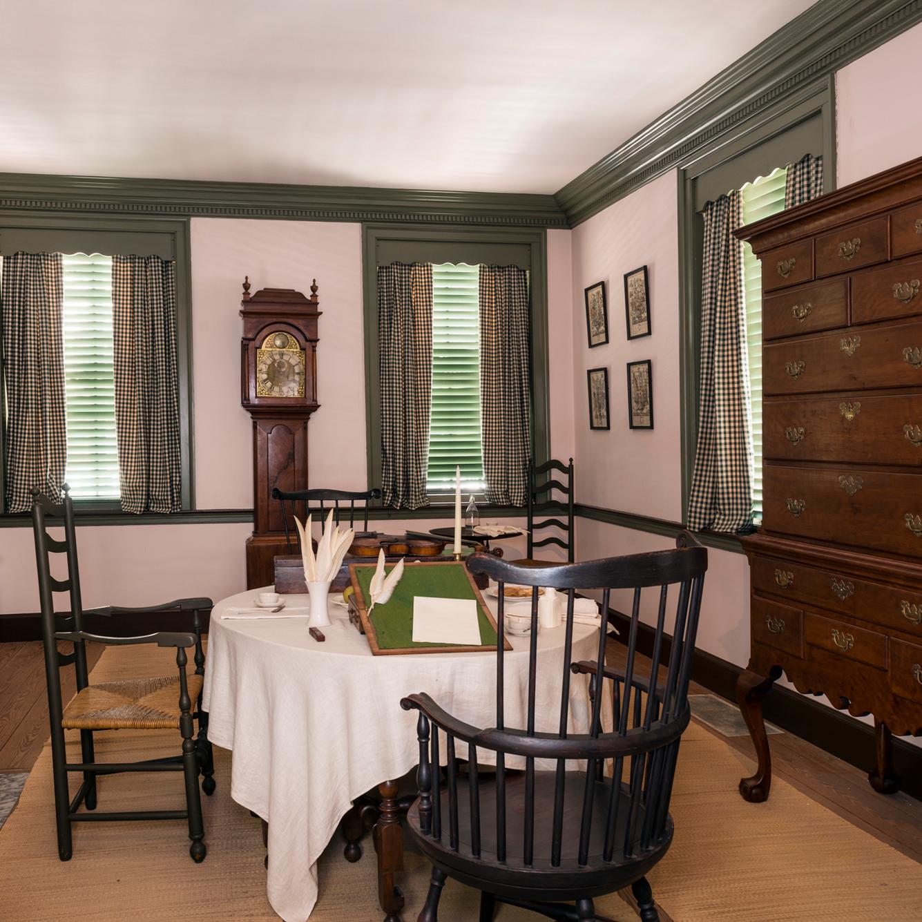 Interior view of a parlor with a swivel chair at a table and a tall case clock along the wall.