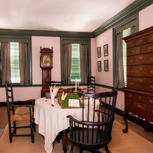 Interior view of a parlor with a swivel chair at a table and a tall case clock along the wall.