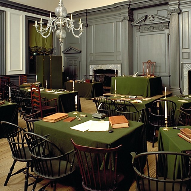 Two rows of t ables and chairs sit in a semi-circle facing a dais with one large at a table.