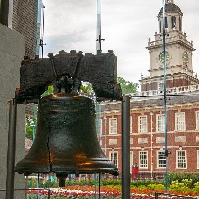 Liberty Bell in the foreground with a large window and viewing Independence Hall