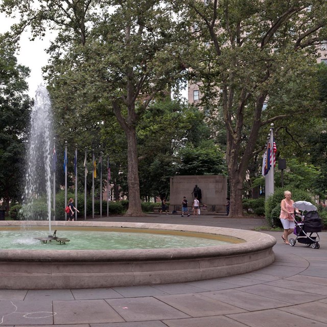 Color photo of an outdoor park setting with fountain in foreground and monument in background.