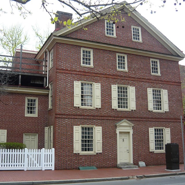 Color photo of two attached red brick buildings; a white fence sits in front of the smaller building