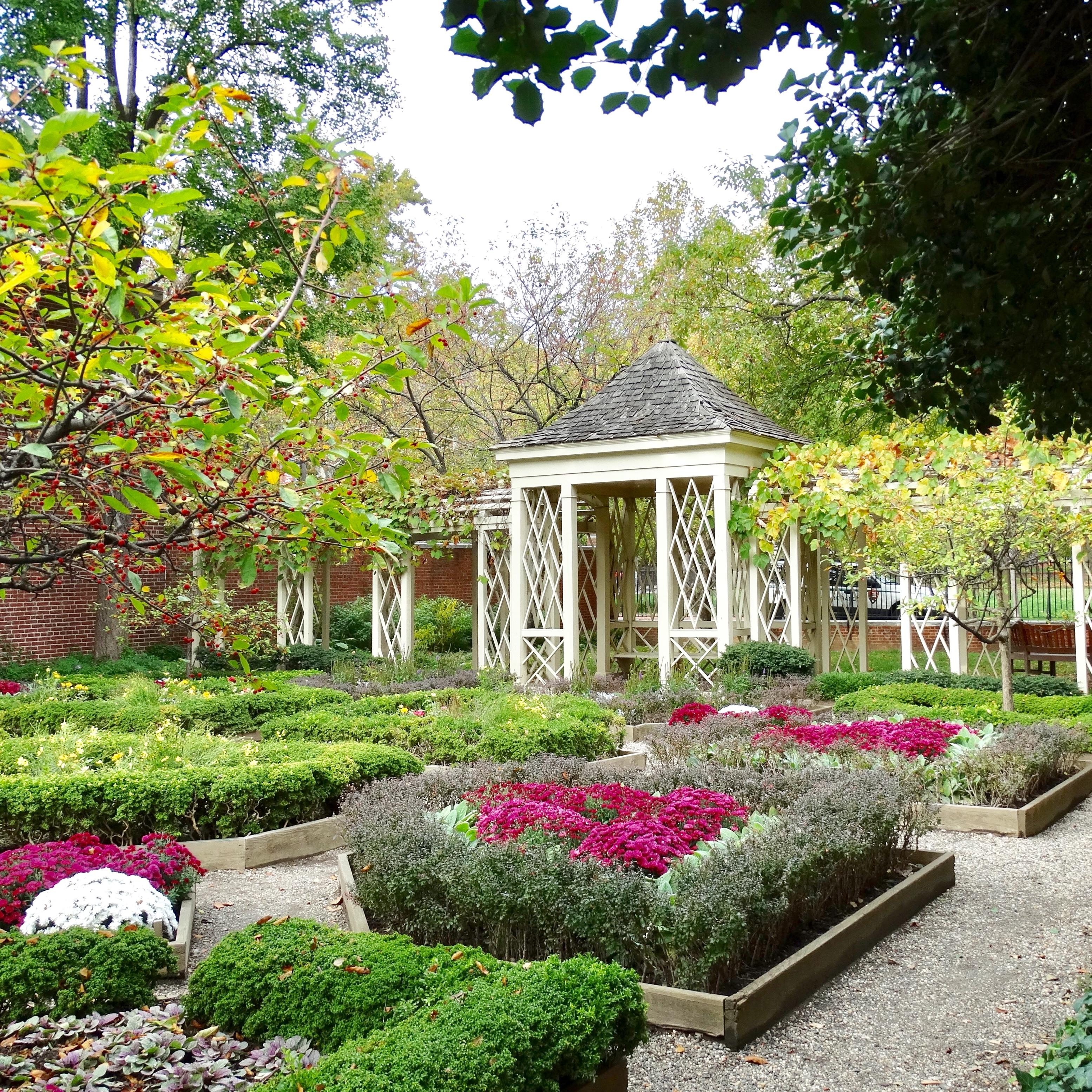 Color photo of gravel path between flowerbeds and trees leading up to white shaded walkway.