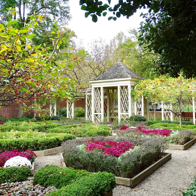 Color photo of gravel path between flowerbeds and trees leading up to white shaded walkway.
