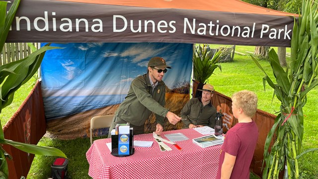 Male NPS rangers under a tent with a table of educational props and brochures 
