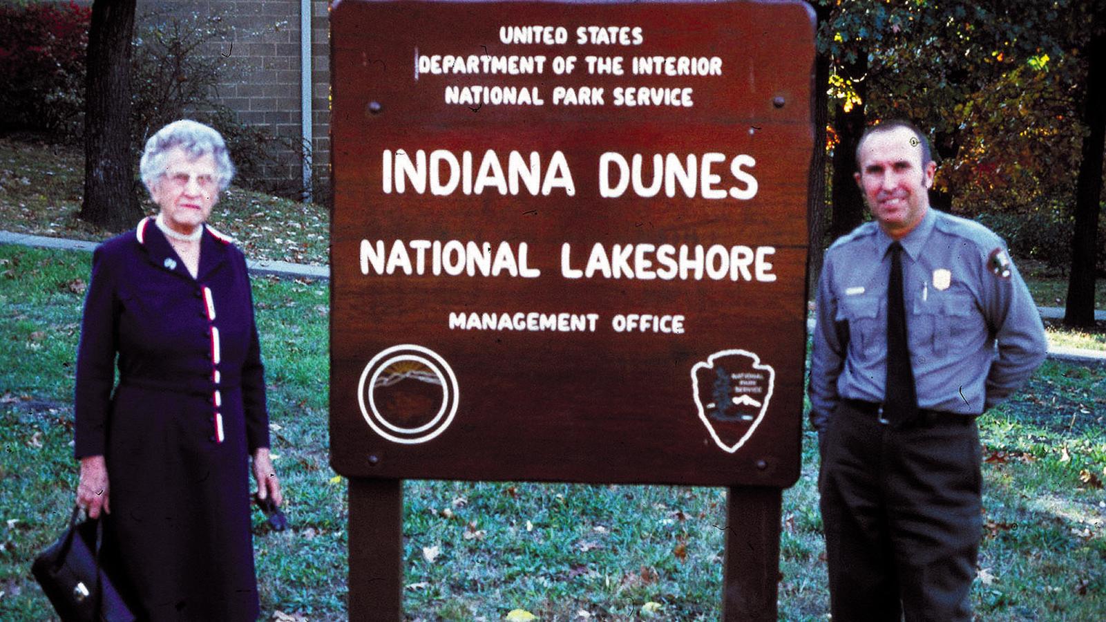 Two individuals stand beside a large &ldquo;Indiana Dunes National Lakeshore&rdquo; sign in an outdoor setting.