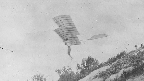 Historic black and white photograph of a man hanging from a bi-wing glider as he glides from a dune.
