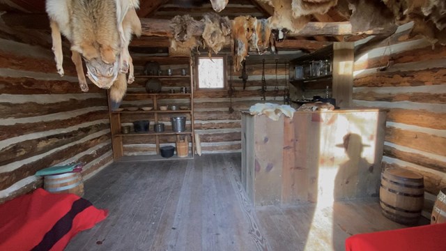 Interior of Bailly log storehouse; rough wooden log walls, animal furs, shelves with cups and bowls