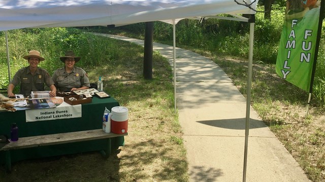 Male and female NPS rangers under a tent with a table of educational props and brochures 