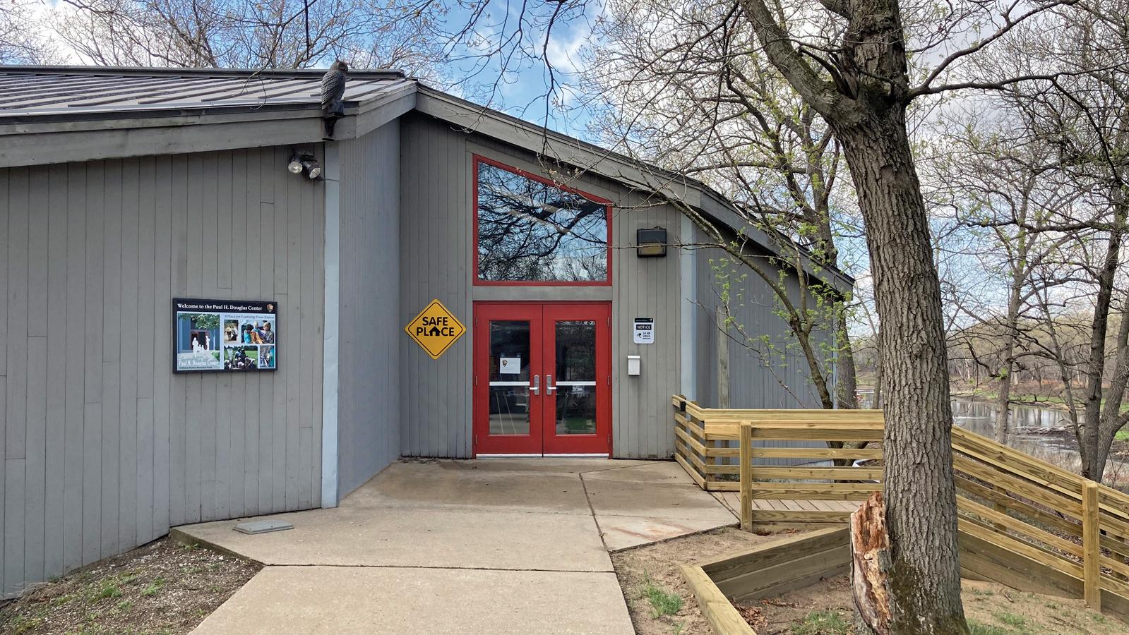Gray building with red doors, Safe Place sign, trees, ramp, and a park setting near a river.