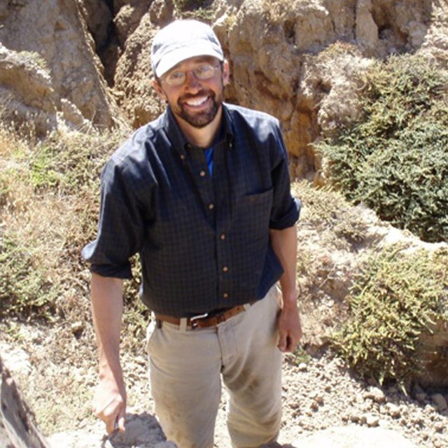Man with blue shirt and white hat standing in rock formations. 