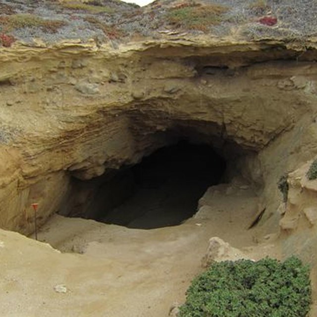 Tan rock with cliffs leading to a dark cave. 