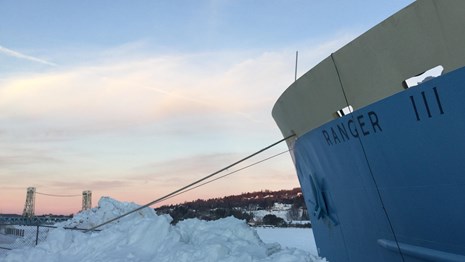 Bow of Ranger III Ferry in winter. Snow around ship. Sunset behind Lift Bridge in distance