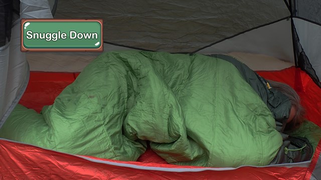 A person huddled in a green sleeping bag inside a red tent.