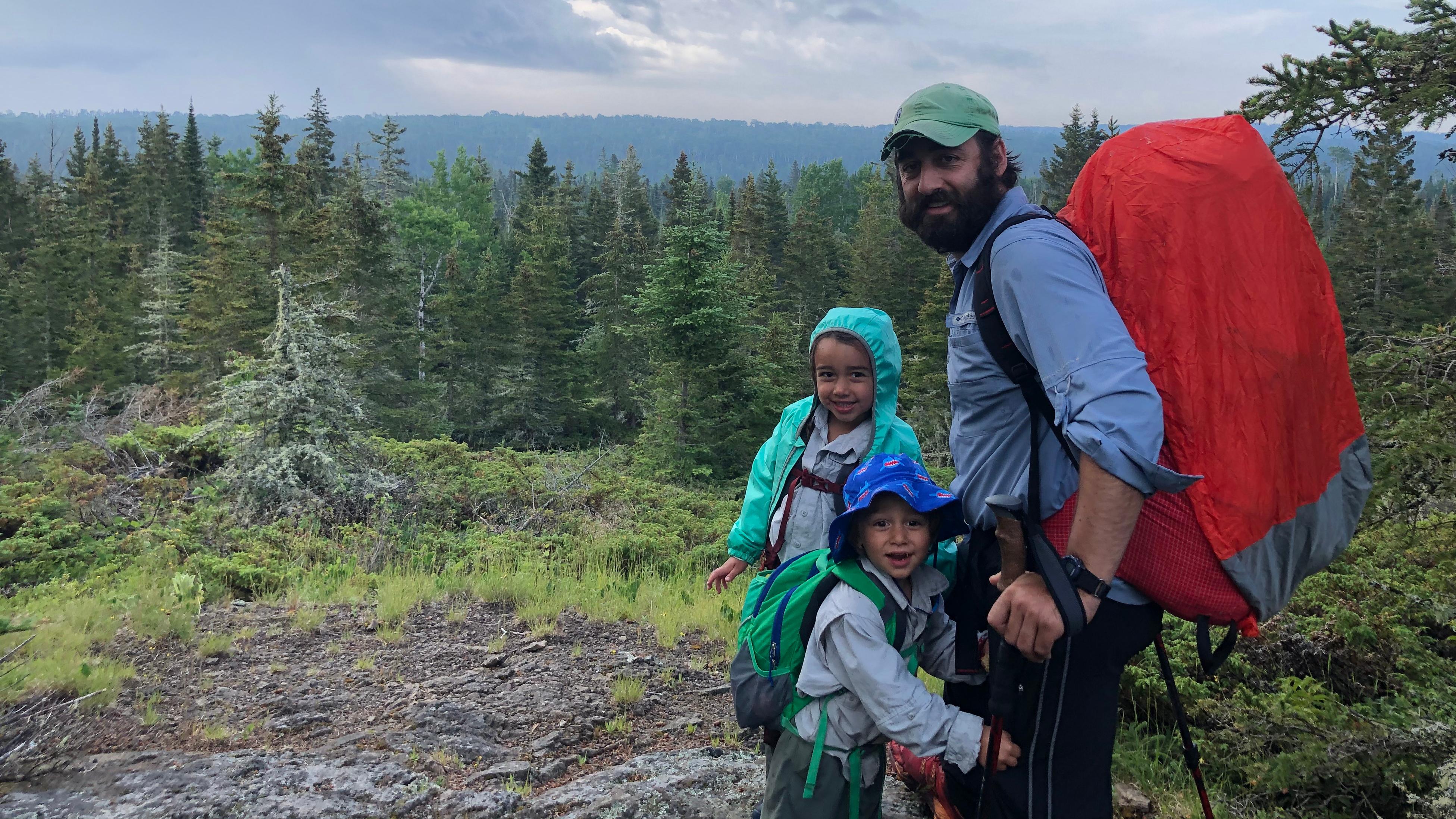 An adult with a backpack on stands next to two children along a rocky trail in a forest.