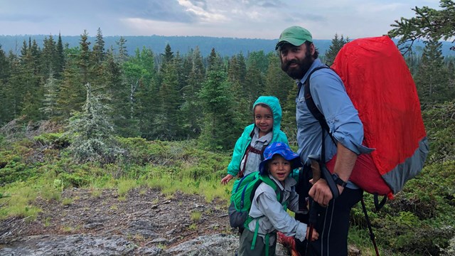 An adult with a backpack on stands next to two children along a rocky trail in a forest.