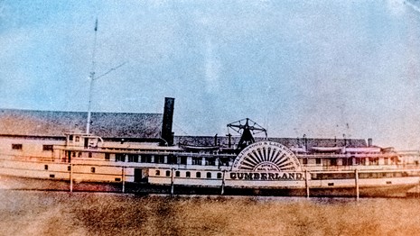 view of SS Cumberland docked with building behind it, large wheel bearing the Cumberland's name