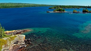 Rugged Isle Royale coastline with blue water, shallow reefs, forest, and rocks.