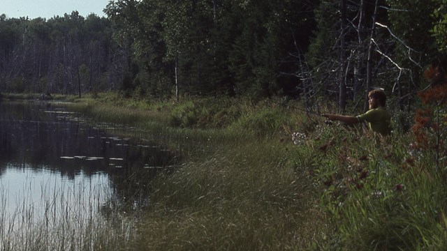 A person stands along a shoreline casting a fishing line into a lake.  