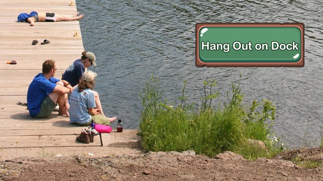 Multiple people relax on a large wooden dock next to a lake.