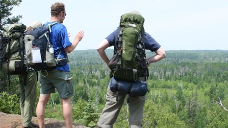 Three people with backpacks stand on ridge overlooking a forest and interior lakes. 