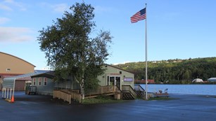 The Houghton Visitor Center building is surrounded by parking lot next to a body of water.
