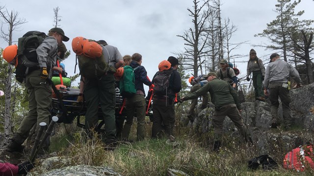A group of park rangers carry an injured hiker on a litter across a rocky ridge.