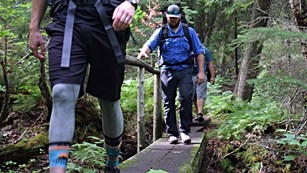 Two people with backpacks on walk across a wooden bridge in the forest.