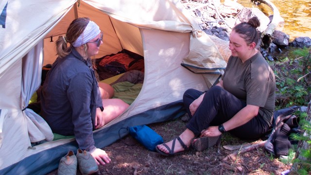 Two people talk while sitting on the ground near a tent.