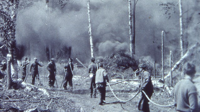 Firefighters at work near a large, smoky fire. They are holding a long fire hose.