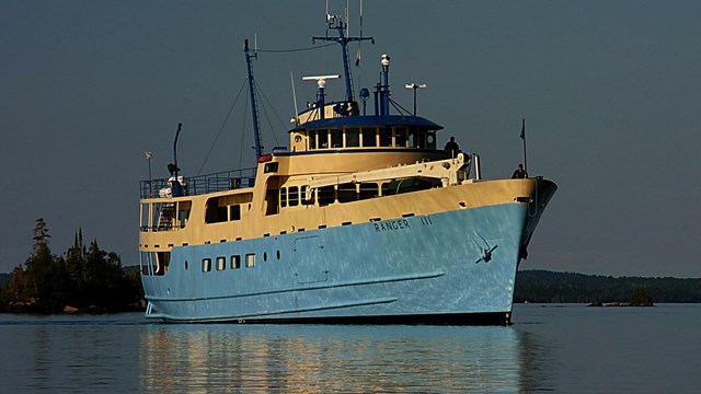 A large blue boat with white upper trim shines in the morning sun with Isle Royale in the background