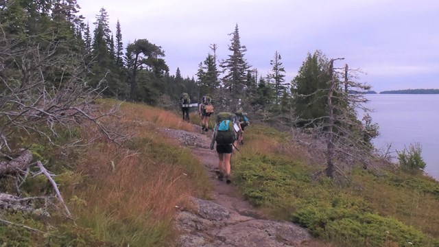 Four people with backpacks hike along a rocky trail next to water with trees in the distance. 